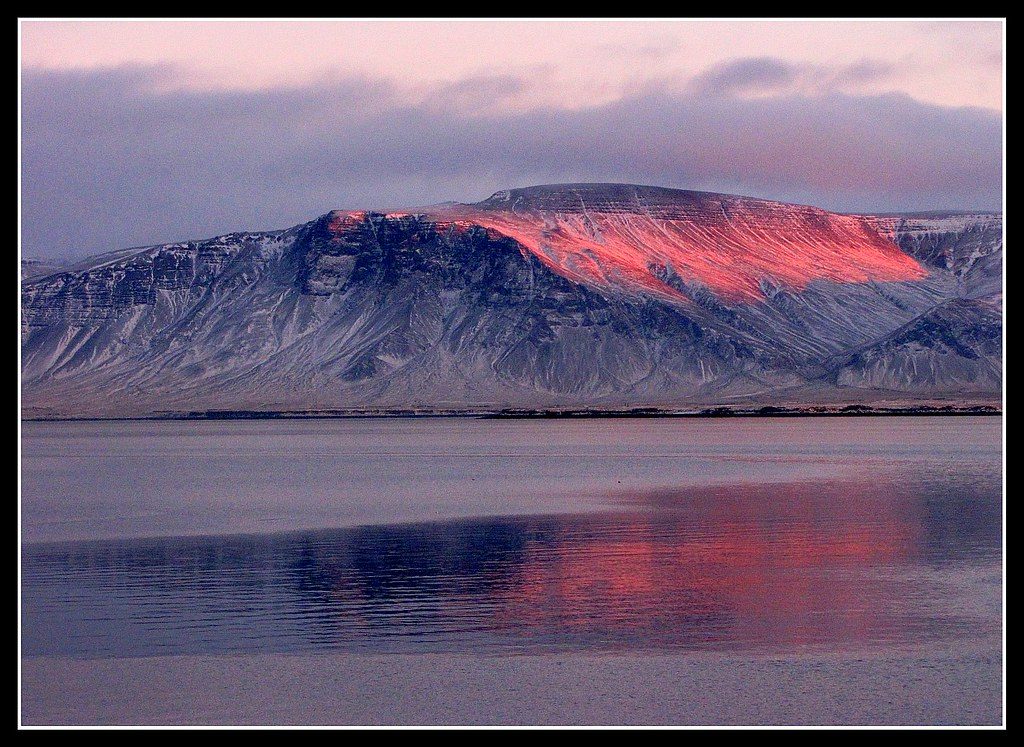 Alpenglow lights the snow-streaked ridge of Mount Esja in Iceland pink-orange at sunset, mirrored in the calm fjord below.
